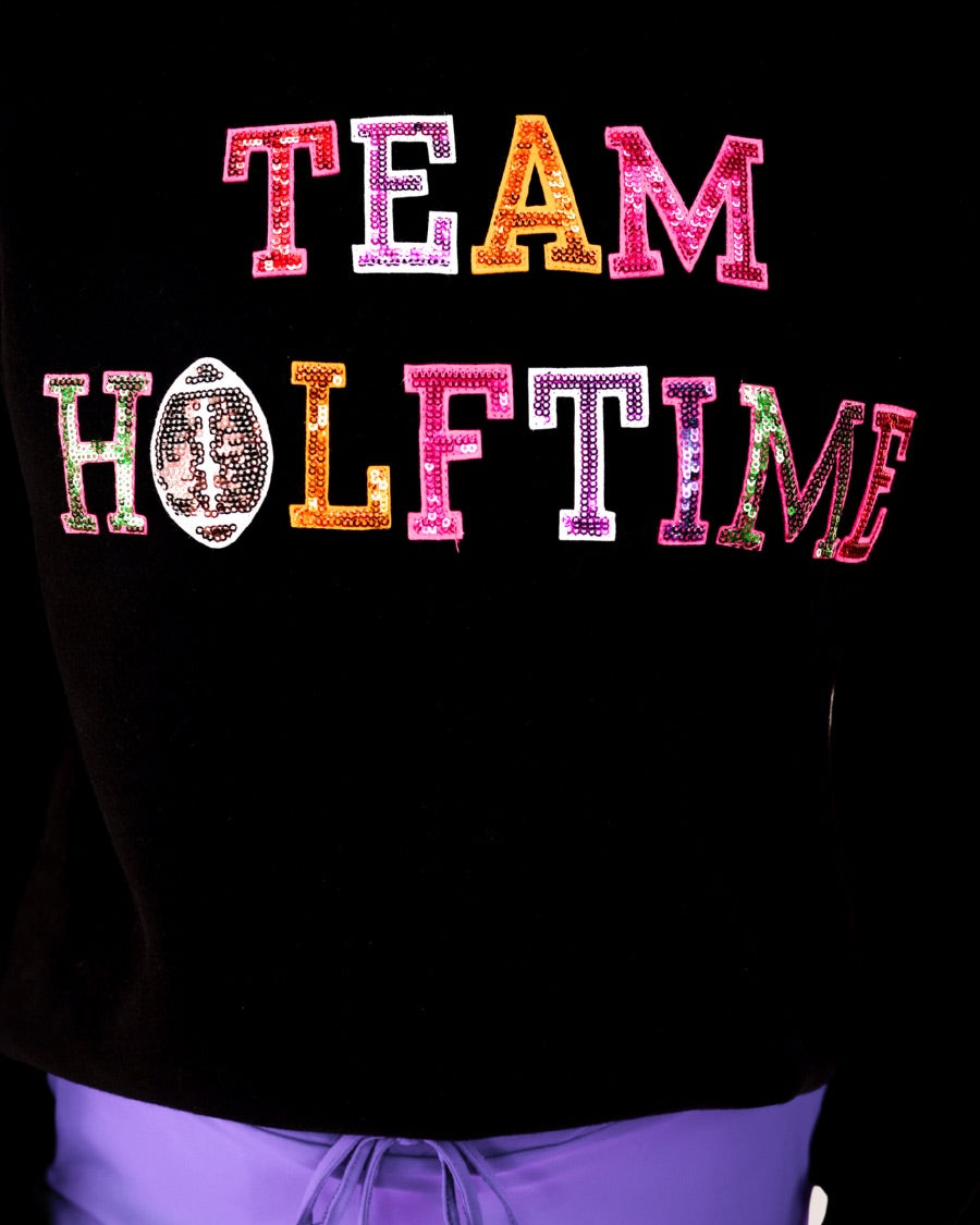 black long-sleeve sweatshirt made from soft mid-weight fabric with a crew neckline, straight hem, and bold multicolored varsity-style chenille patches that spell out 'TEAM HALFTIME' across the front chest