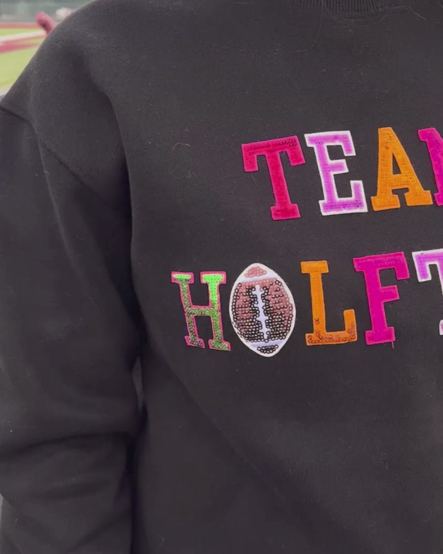 black long-sleeve sweatshirt made from soft mid-weight fabric with a crew neckline, straight hem, and bold multicolored varsity-style chenille patches that spell out 'TEAM HALFTIME' across the front chest