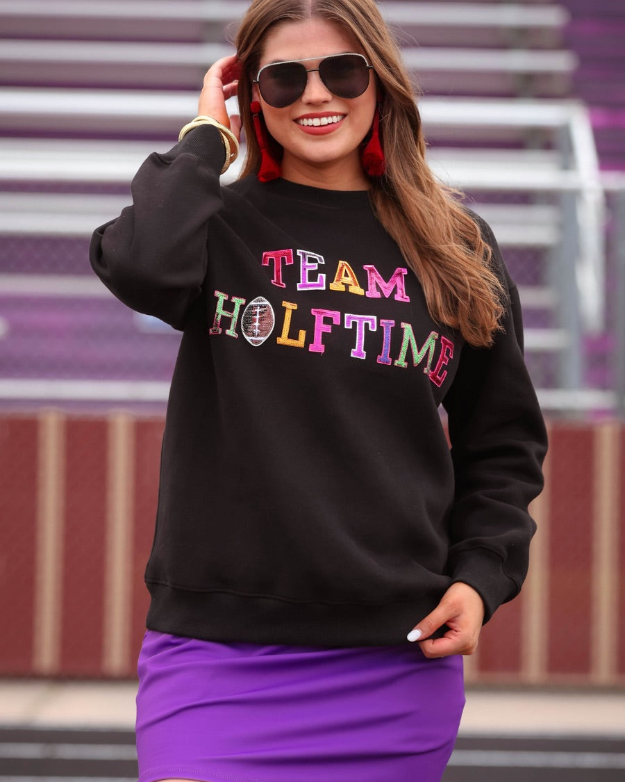 black long-sleeve sweatshirt made from soft mid-weight fabric with a crew neckline, straight hem, and bold multicolored varsity-style chenille patches that spell out 'TEAM HALFTIME' across the front chest