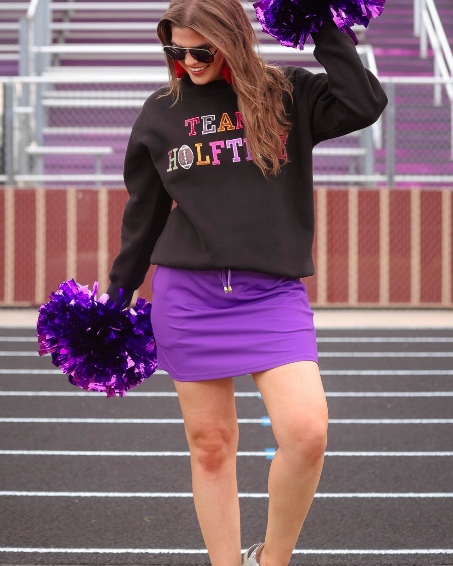 black long-sleeve sweatshirt made from soft mid-weight fabric with a crew neckline, straight hem, and bold multicolored varsity-style chenille patches that spell out 'TEAM HALFTIME' across the front chest