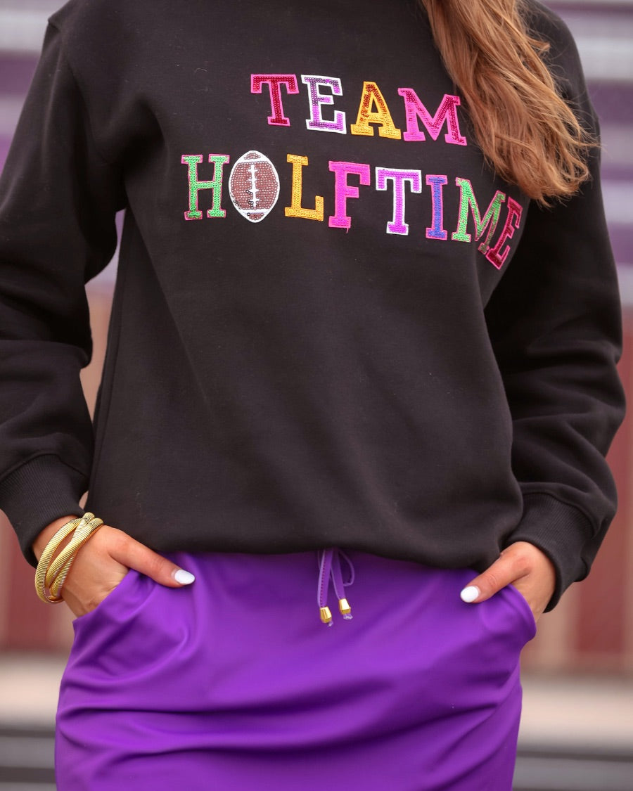 black long-sleeve sweatshirt made from soft mid-weight fabric with a crew neckline, straight hem, and bold multicolored varsity-style chenille patches that spell out 'TEAM HALFTIME' across the front chest