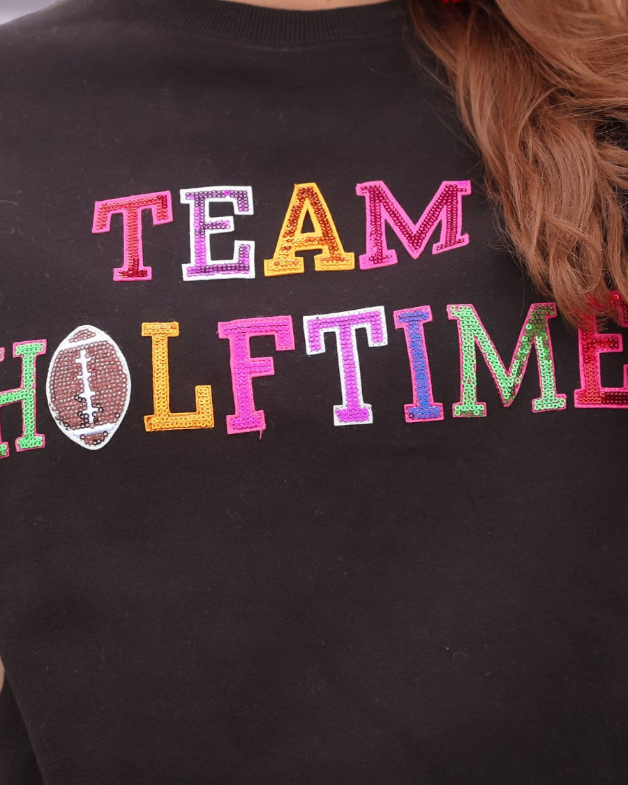 black long-sleeve sweatshirt made from soft mid-weight fabric with a crew neckline, straight hem, and bold multicolored varsity-style chenille patches that spell out 'TEAM HALFTIME' across the front chest