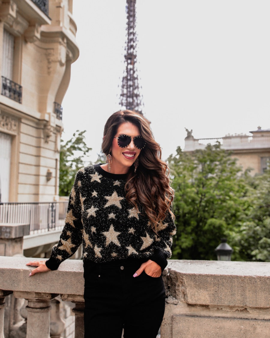 a black long-sleeve crewneck sweater with a festive star motif, shimmering tinsel woven throughout, and ribbed trim at the neckline, cuffs, and hemline