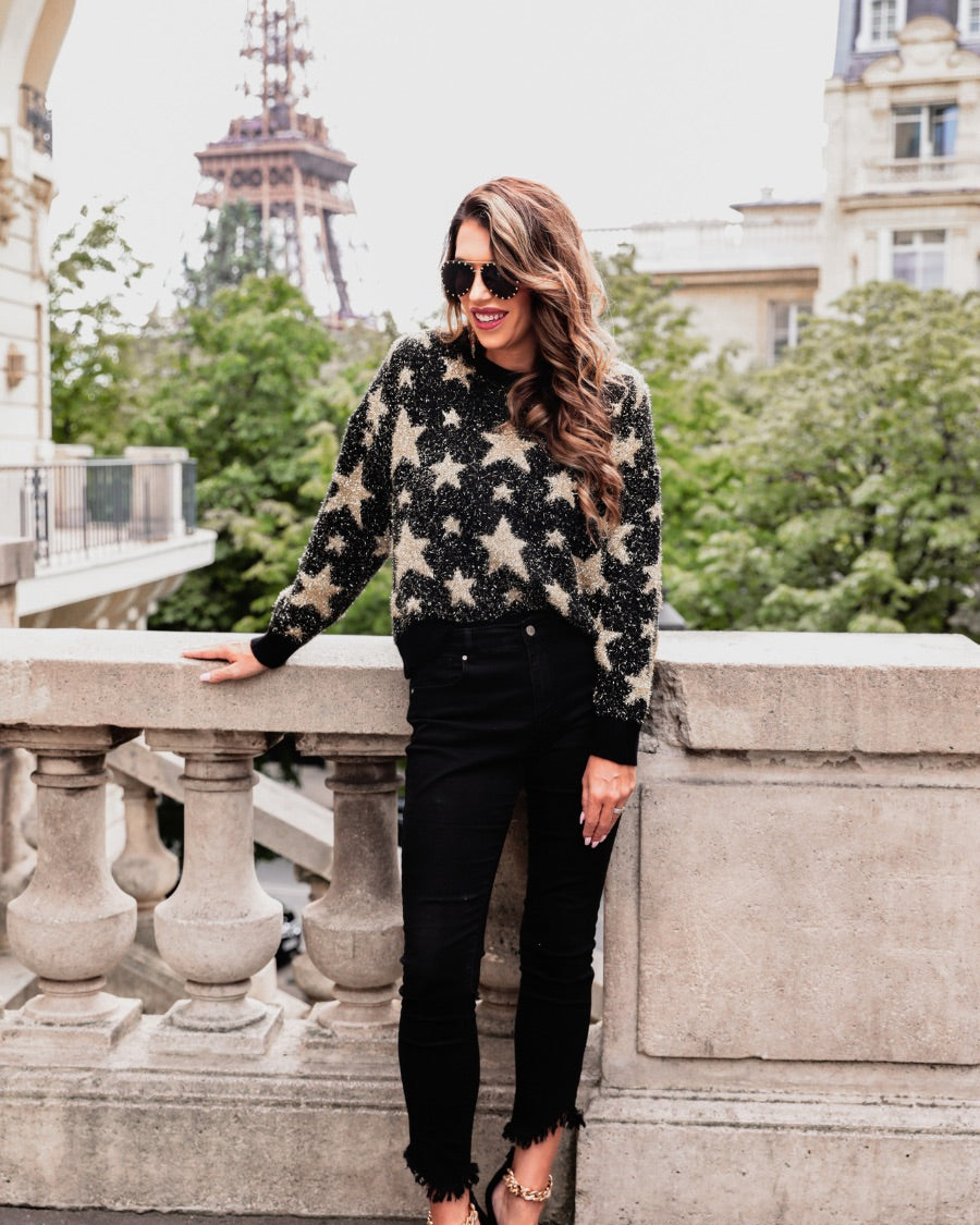 a black long-sleeve crewneck sweater with a festive star motif, shimmering tinsel woven throughout, and ribbed trim at the neckline, cuffs, and hemline