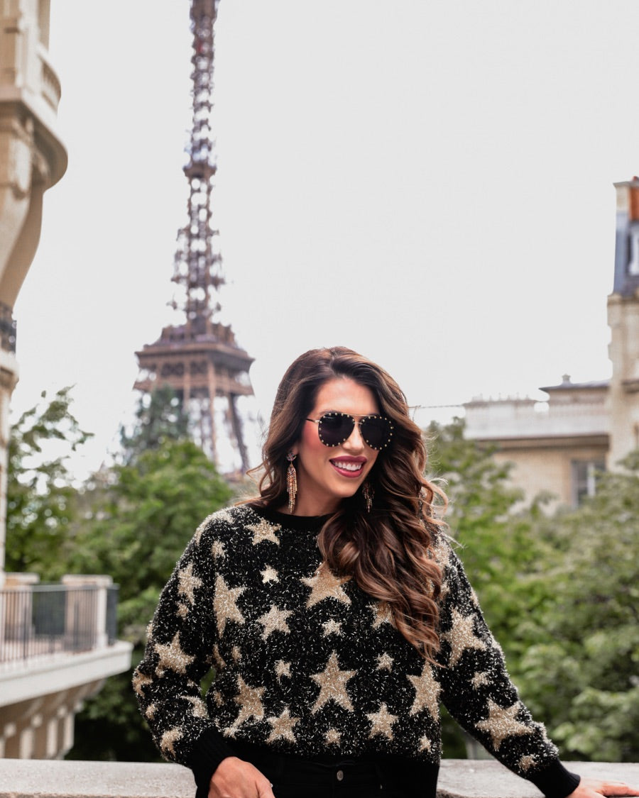 a black long-sleeve crewneck sweater with a festive star motif, shimmering tinsel woven throughout, and ribbed trim at the neckline, cuffs, and hemline