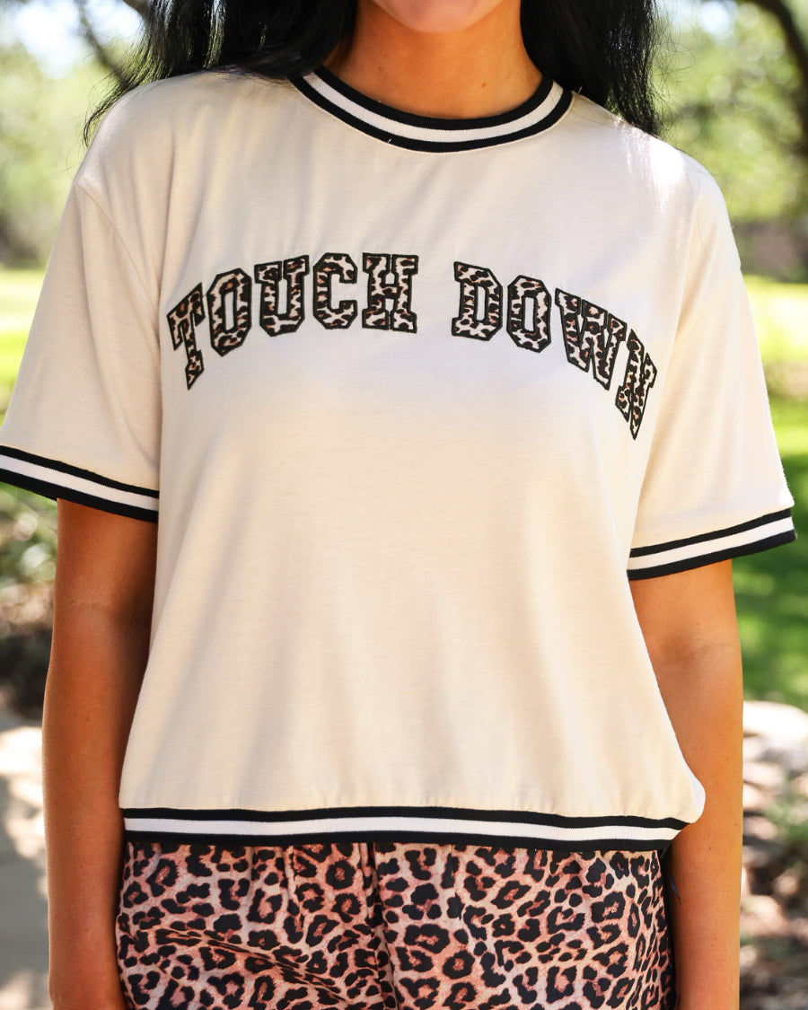 a boxy fit crew neck tee with a leopard 'TOUCH DOWN' fabric patch, embroidered outline, contrast neckline and sleeve trim, short sleeves, and soft stretch fabric on a cream base