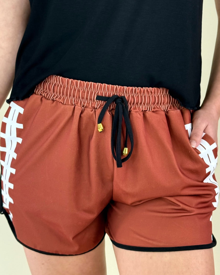 mid-rise relaxed fit activewear shorts with side pockets and a tie waist drawstring with gold bead accent, designed with a football-lace inspired pattern print fabric down each leg, brown base, and black trim