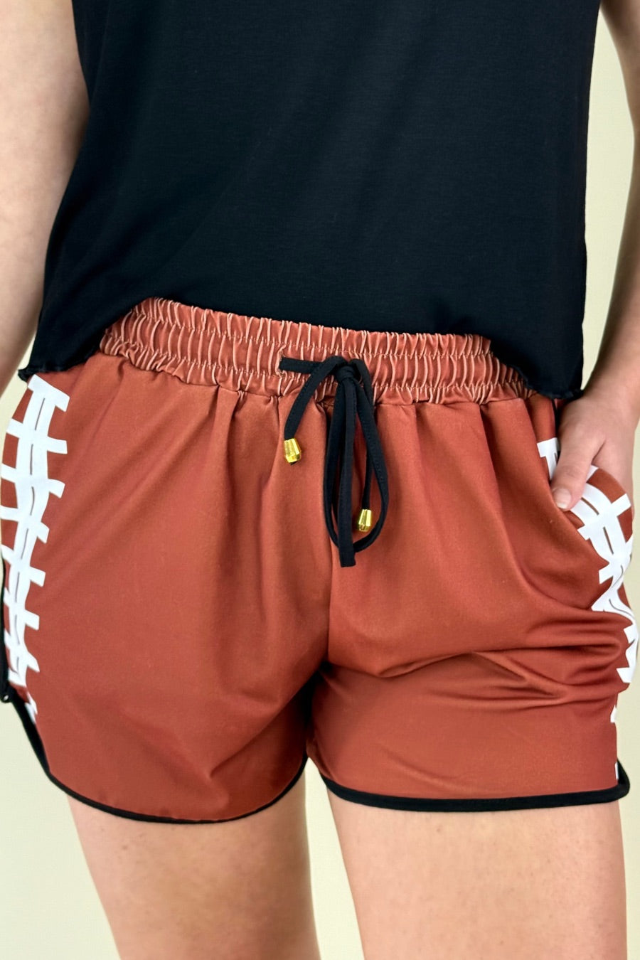 mid-rise relaxed fit activewear shorts with side pockets and a tie waist drawstring with gold bead accent, designed with a football-lace inspired pattern print fabric down each leg, brown base, and black trim