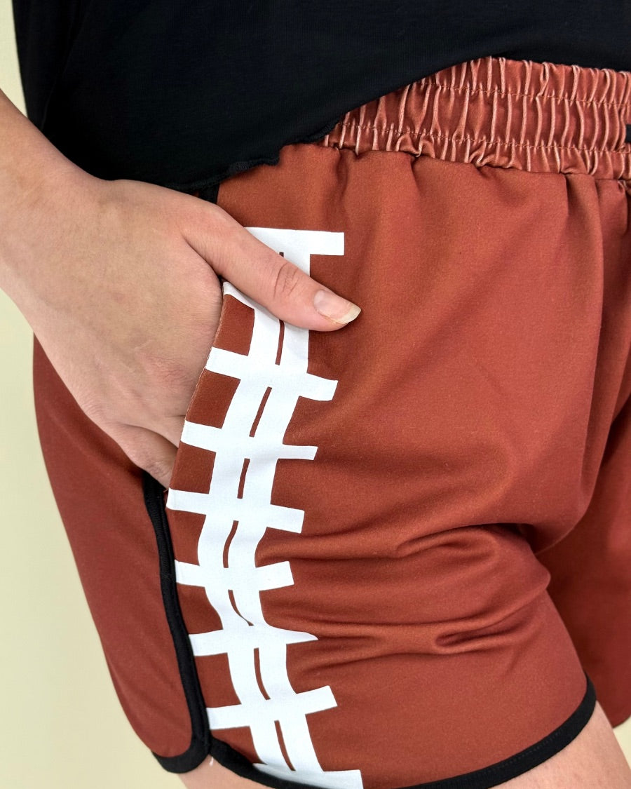 mid-rise relaxed fit activewear shorts with side pockets and a tie waist drawstring with gold bead accent, designed with a football-lace inspired pattern print fabric down each leg, brown base, and black trim