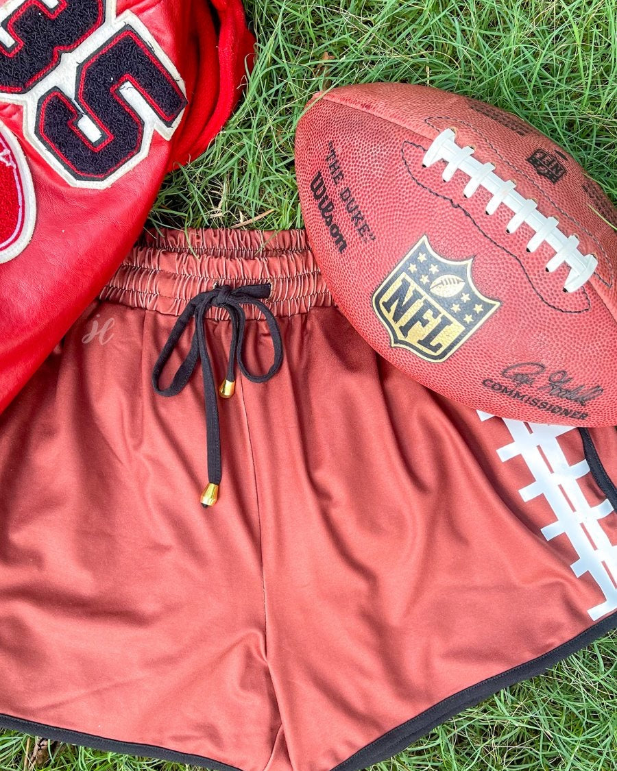 mid-rise relaxed fit activewear shorts with side pockets and a tie waist drawstring with gold bead accent, designed with a football-lace inspired pattern print fabric down each leg, brown base, and black trim