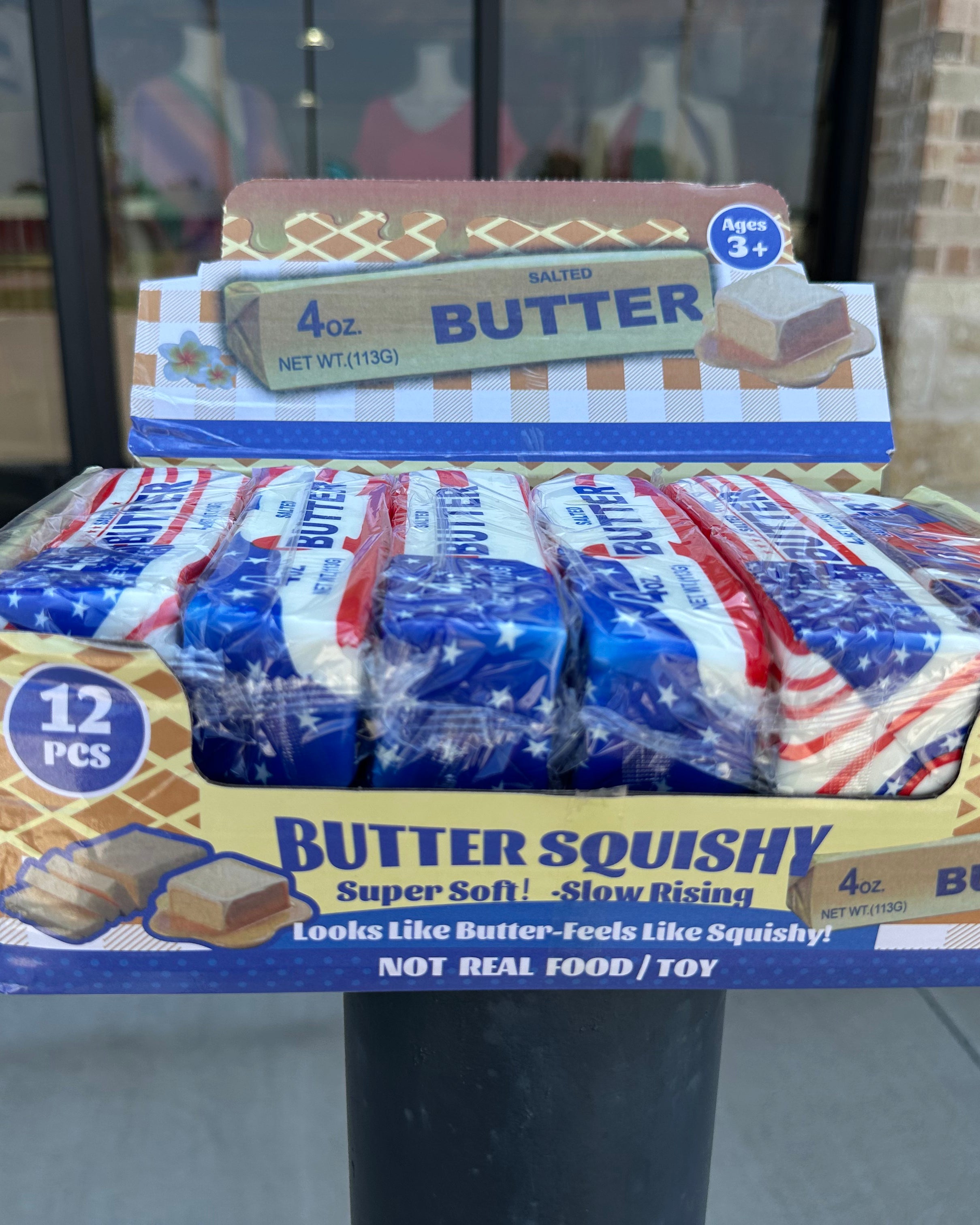 A retail display box filled with individually wrapped Butter Squishy toys, with a smaller display box placed behind it. The packaging features an American flag design with red, white, and blue, and text reading 'BUTTER SQUISHY' and '12 PCS'.