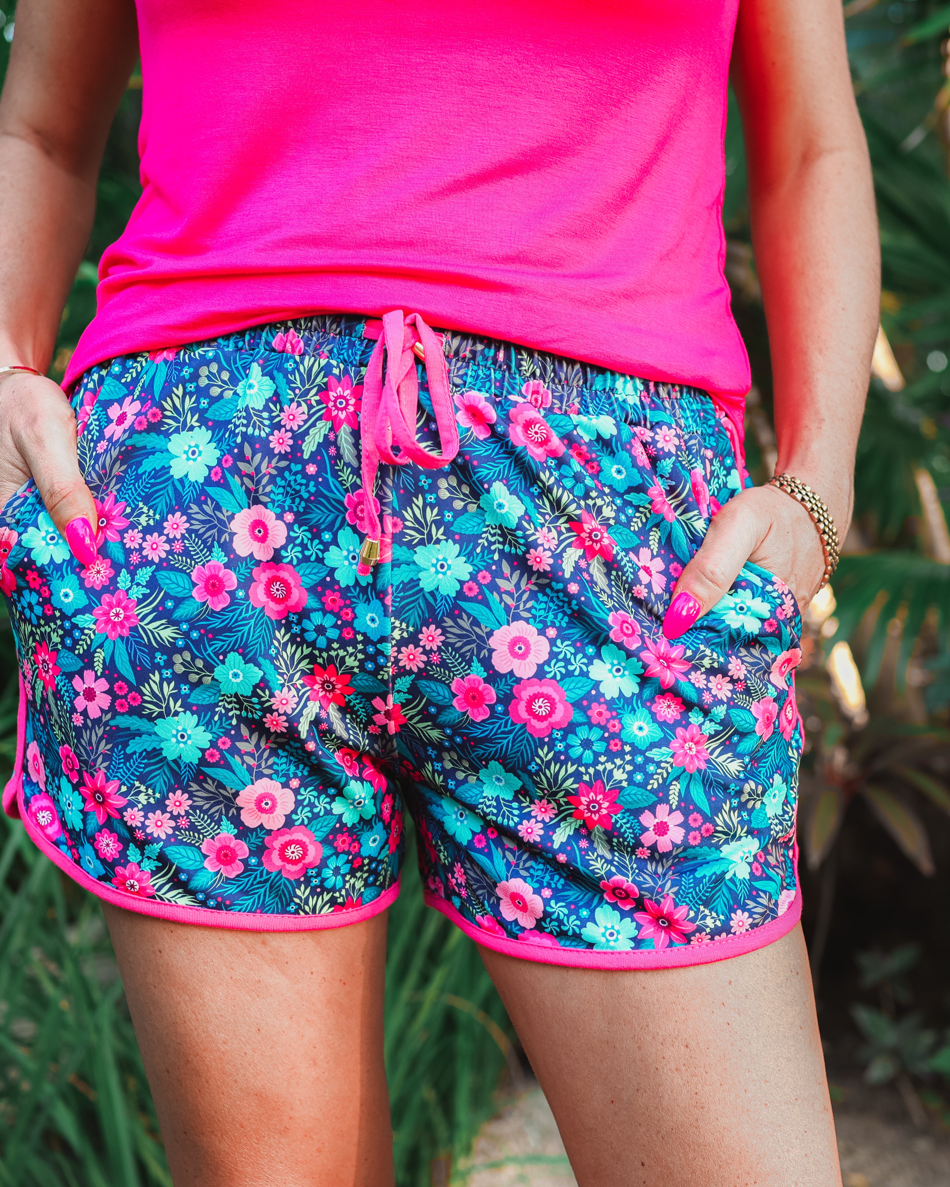 mid-rise relaxed fit activewear shorts with side pockets and a tie waist drawstring with gold bead accents, designed on a vibrant multicolor floral pattern print fabric, navy base, and pink trim