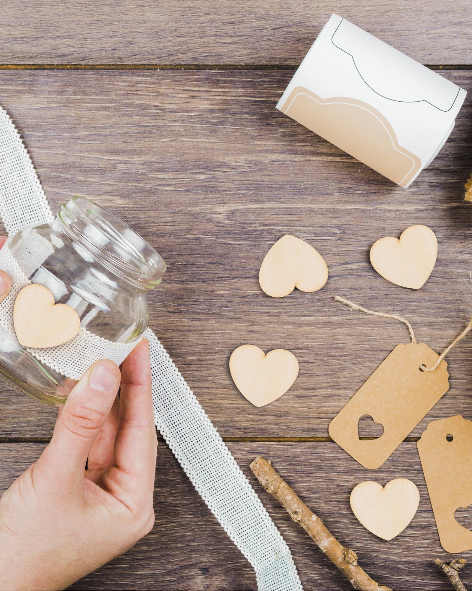 Hand holding a glass jar with heart-shaped decorations on a wooden surface with ribbons and tags.