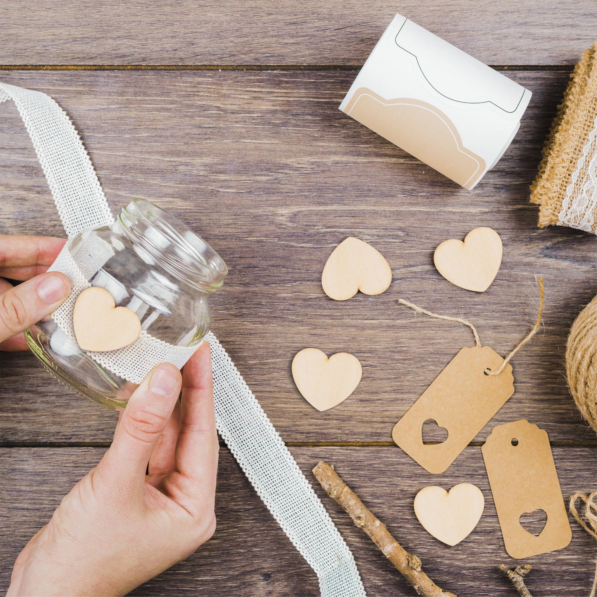 Hand holding a glass jar with heart-shaped decorations on a wooden surface with ribbons and tags.