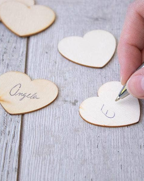 Hand writing on a heart-shaped wooden tag with other heart-shaped tags in the background.
