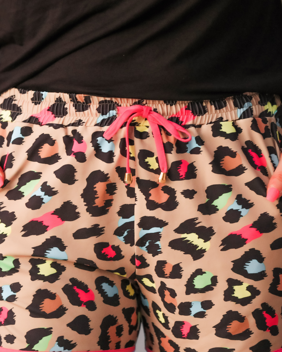 mid-rise relaxed fit activewear shorts with side pockets and a tie waist drawstring with gold bead accents, designed on a vibrant multicolor leopard pattern print fabric, tan base, and pink trim