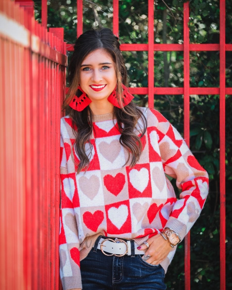 a long-sleeve crew neckline sweater with a relaxed drop-shoulder silhouette and ribbed hemline, neckline, and cuffs, plus a contrasting color block heart pattern in ivory, red, and taupe