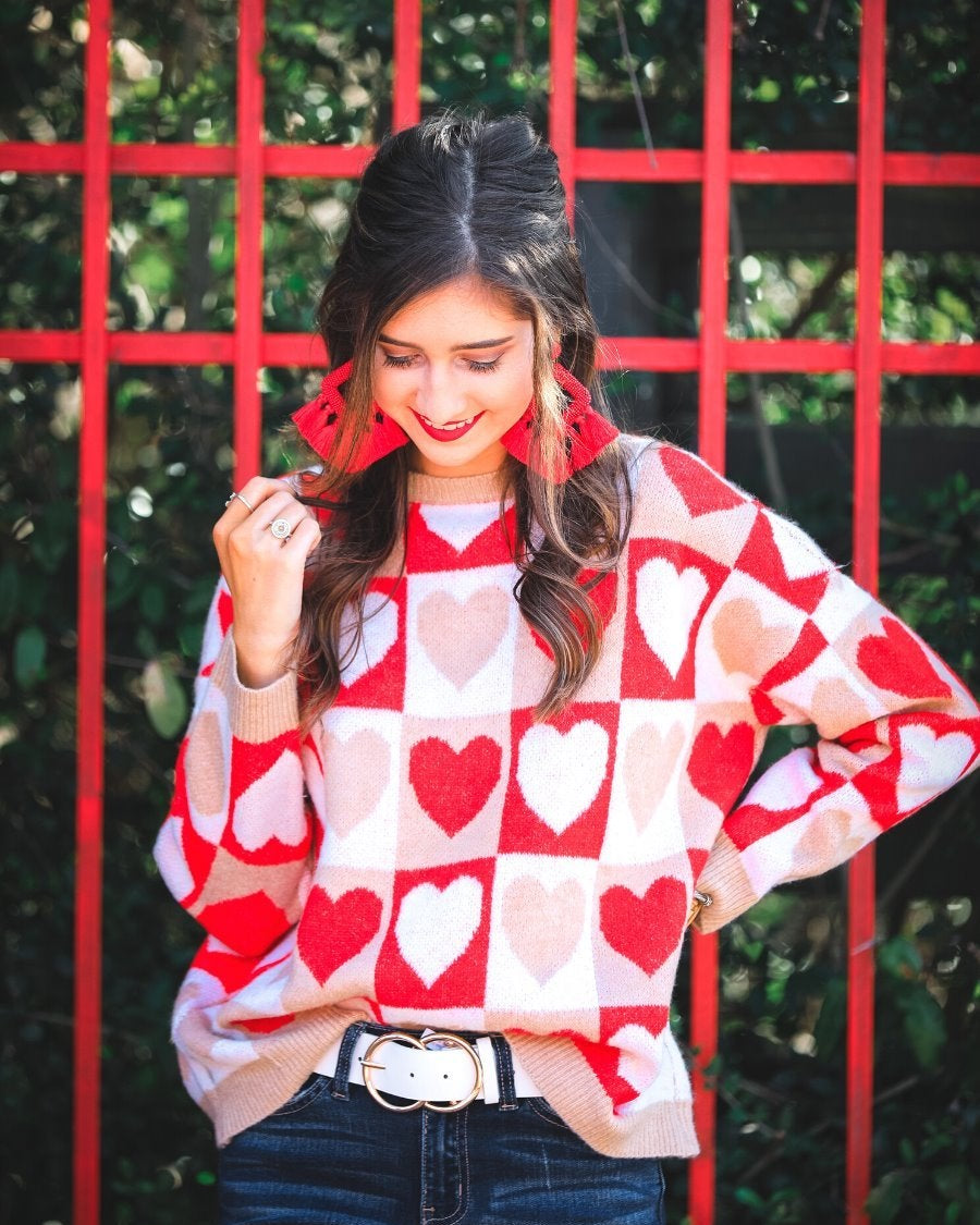 a long-sleeve crew neckline sweater with a relaxed drop-shoulder silhouette and ribbed hemline, neckline, and cuffs, plus a contrasting color block heart pattern in ivory, red, and taupe