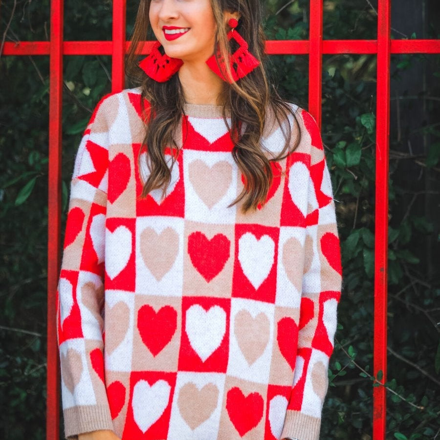 a long-sleeve crew neckline sweater with a relaxed drop-shoulder silhouette and ribbed hemline, neckline, and cuffs, plus a contrasting color block heart pattern in ivory, red, and taupe