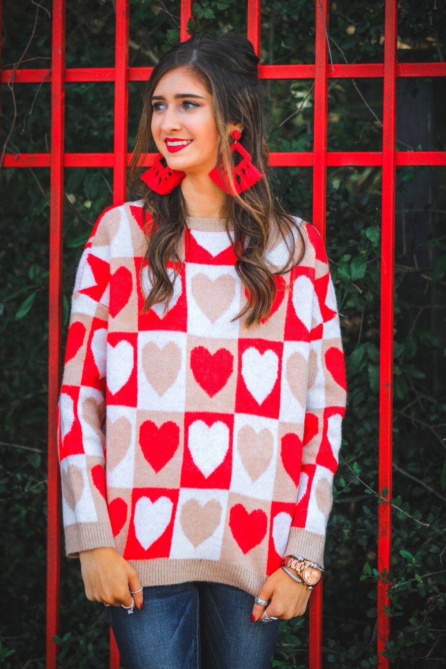 a long-sleeve crew neckline sweater with a relaxed drop-shoulder silhouette and ribbed hemline, neckline, and cuffs, plus a contrasting color block heart pattern in ivory, red, and taupe