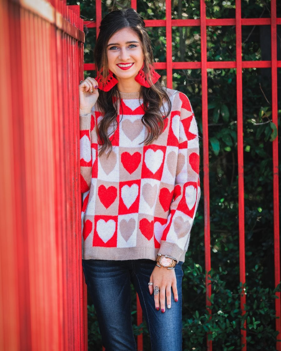 a long-sleeve crew neckline sweater with a relaxed drop-shoulder silhouette and ribbed hemline, neckline, and cuffs, plus a contrasting color block heart pattern in ivory, red, and taupe