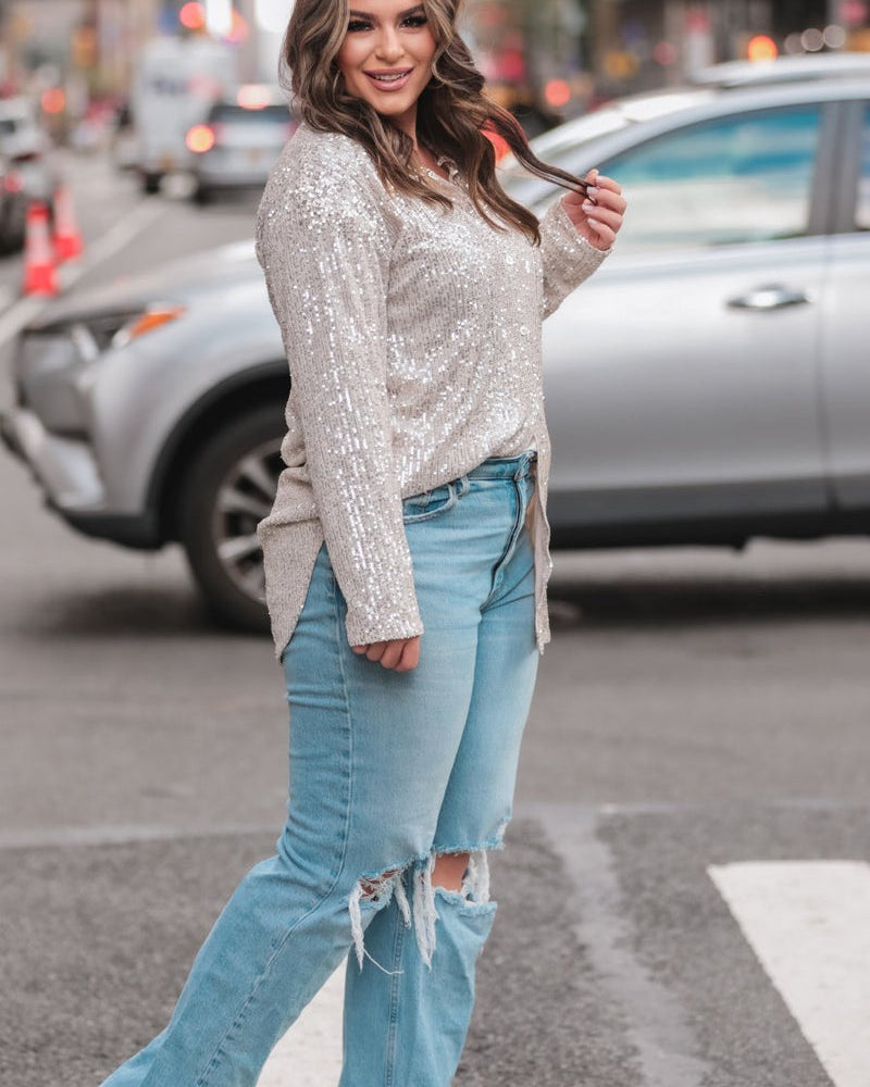 a loose long-sleeve top made of lightweight material covered in shimmering sequins with a collared neckline, fitted button closure cuffs, an accent chest pocket, and a relaxed button-up silhouette that falls into a round high-low hemline in champagne