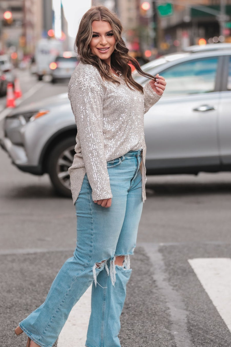 a loose long-sleeve top made of lightweight material covered in shimmering sequins with a collared neckline, fitted button closure cuffs, an accent chest pocket, and a relaxed button-up silhouette that falls into a round high-low hemline in champagne