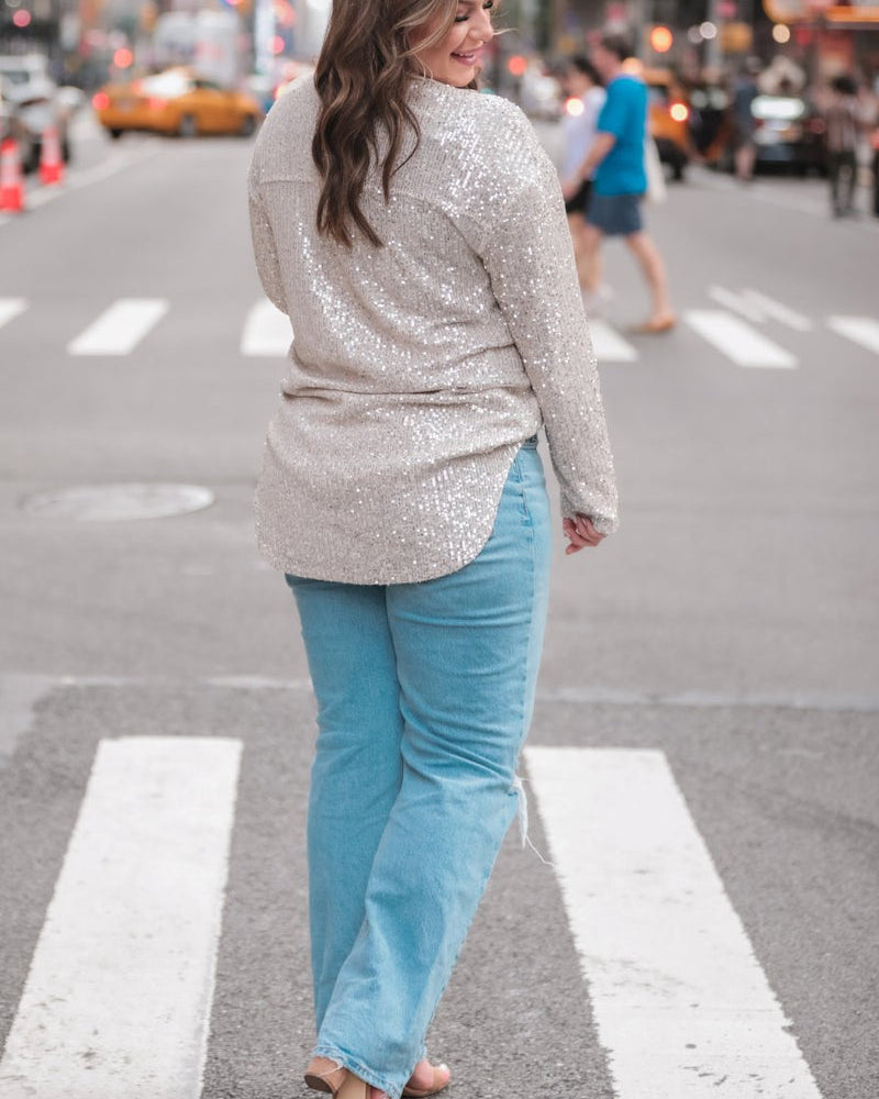 a loose long-sleeve top made of lightweight material covered in shimmering sequins with a collared neckline, fitted button closure cuffs, an accent chest pocket, and a relaxed button-up silhouette that falls into a round high-low hemline in champagne