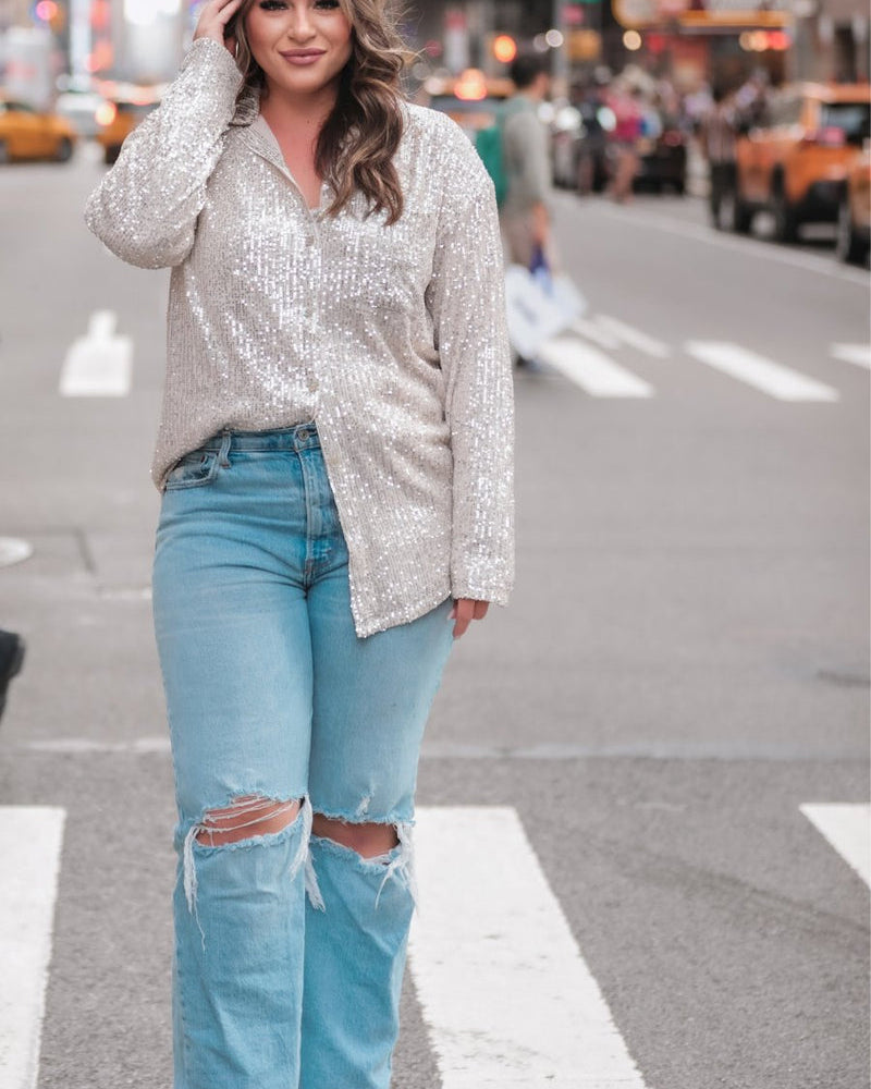 a loose long-sleeve top made of lightweight material covered in shimmering sequins with a collared neckline, fitted button closure cuffs, an accent chest pocket, and a relaxed button-up silhouette that falls into a round high-low hemline in champagne
