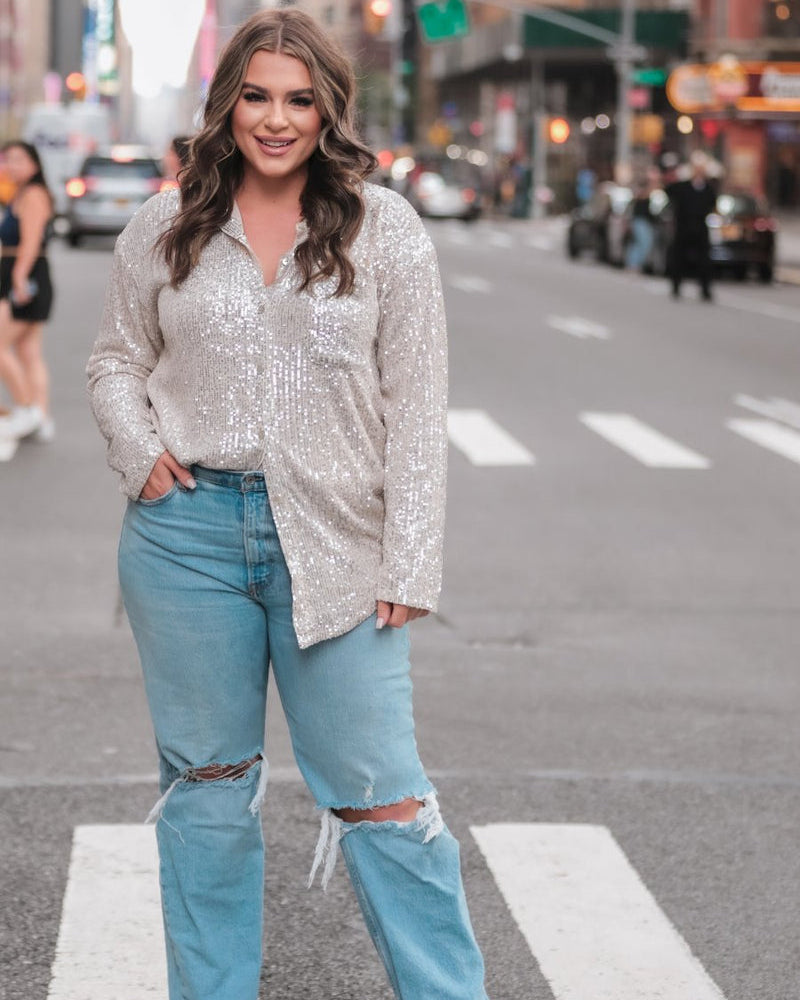 a loose long-sleeve top made of lightweight material covered in shimmering sequins with a collared neckline, fitted button closure cuffs, an accent chest pocket, and a relaxed button-up silhouette that falls into a round high-low hemline in champagne