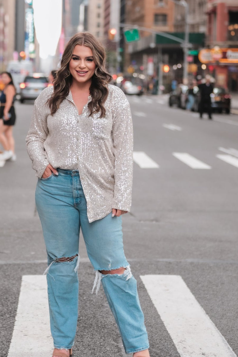 a loose long-sleeve top made of lightweight material covered in shimmering sequins with a collared neckline, fitted button closure cuffs, an accent chest pocket, and a relaxed button-up silhouette that falls into a round high-low hemline in champagne