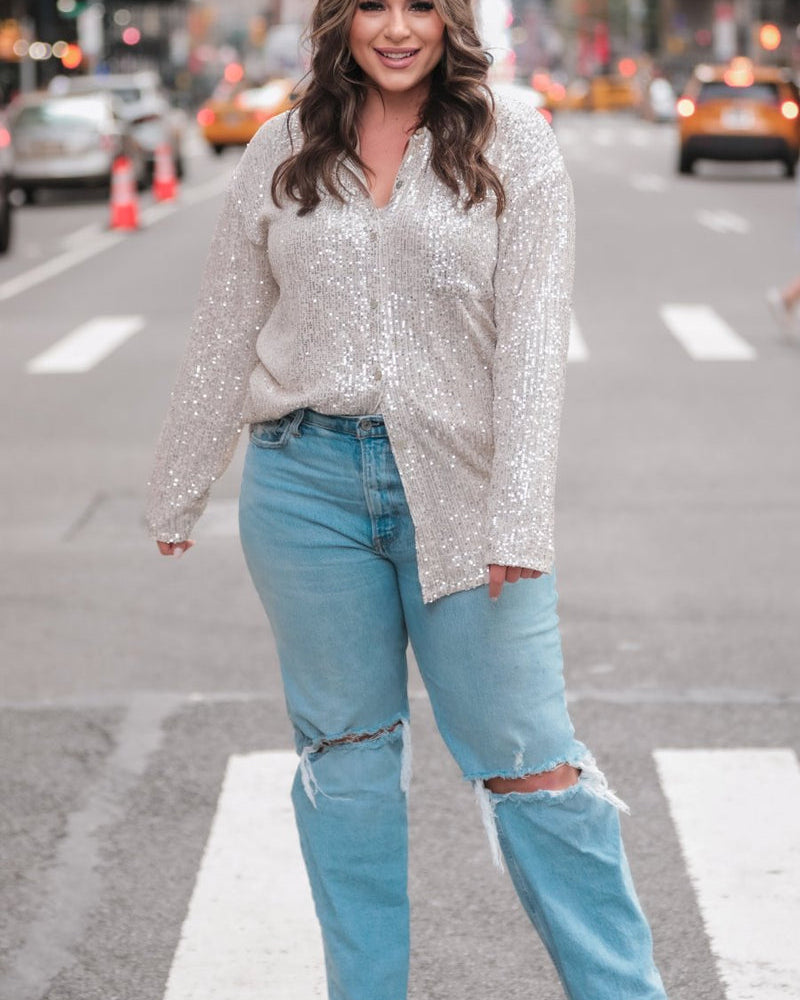 a loose long-sleeve top made of lightweight material covered in shimmering sequins with a collared neckline, fitted button closure cuffs, an accent chest pocket, and a relaxed button-up silhouette that falls into a round high-low hemline in champagne