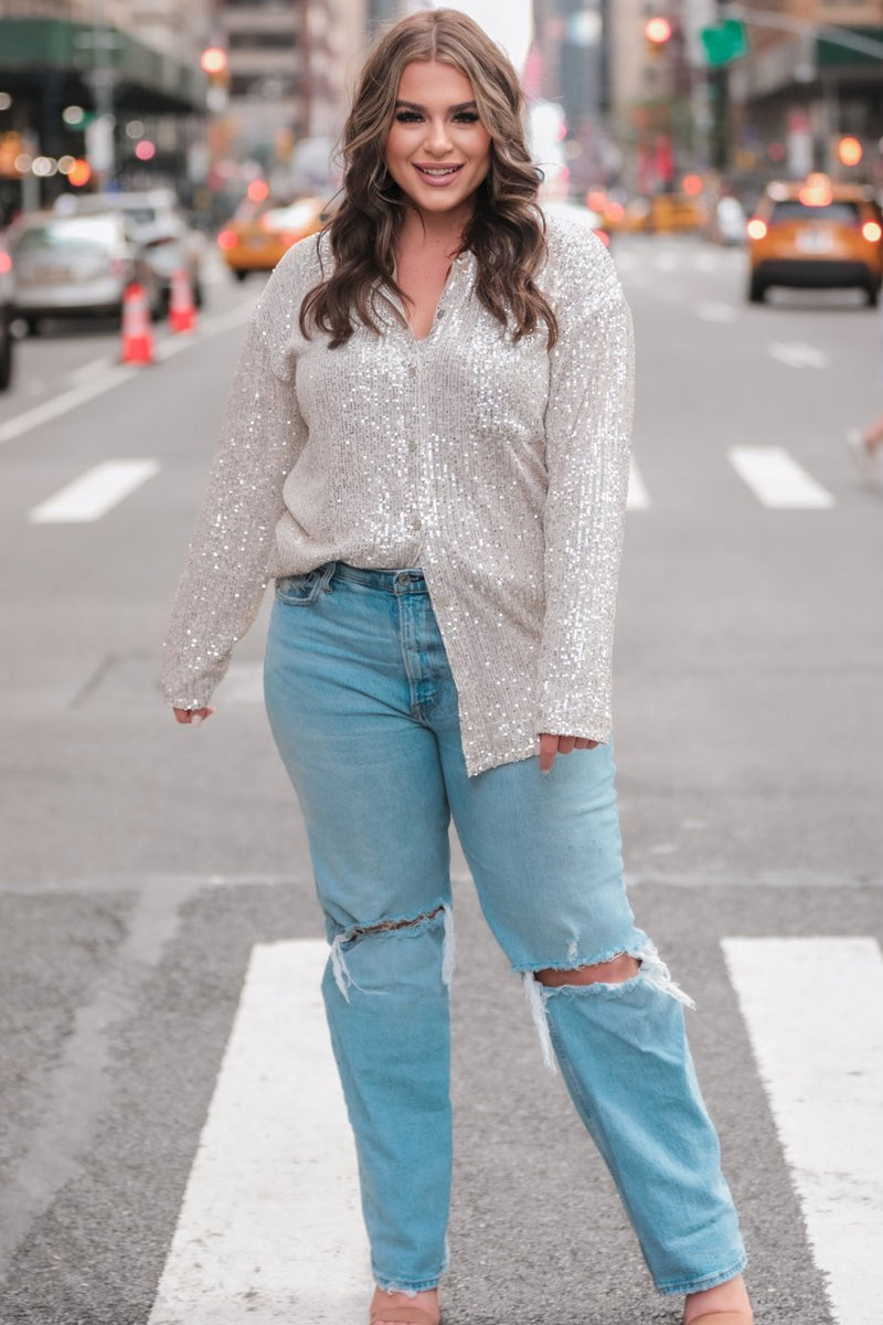 a loose long-sleeve top made of lightweight material covered in shimmering sequins with a collared neckline, fitted button closure cuffs, an accent chest pocket, and a relaxed button-up silhouette that falls into a round high-low hemline in champagne