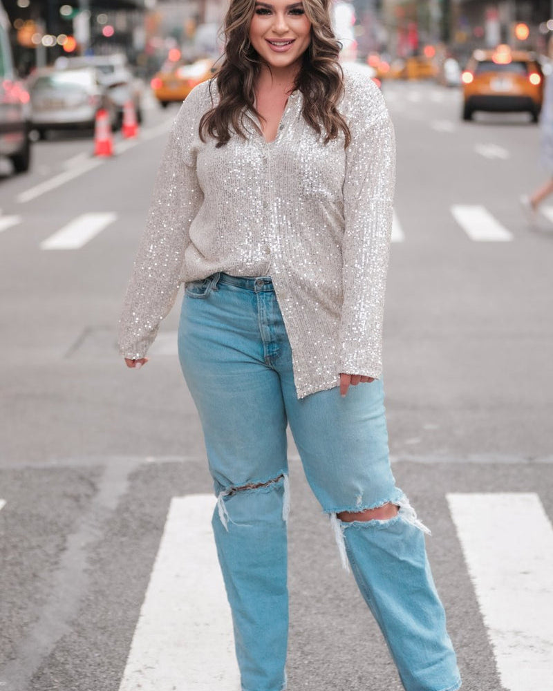 a loose long-sleeve top made of lightweight material covered in shimmering sequins with a collared neckline, fitted button closure cuffs, an accent chest pocket, and a relaxed button-up silhouette that falls into a round high-low hemline in champagne