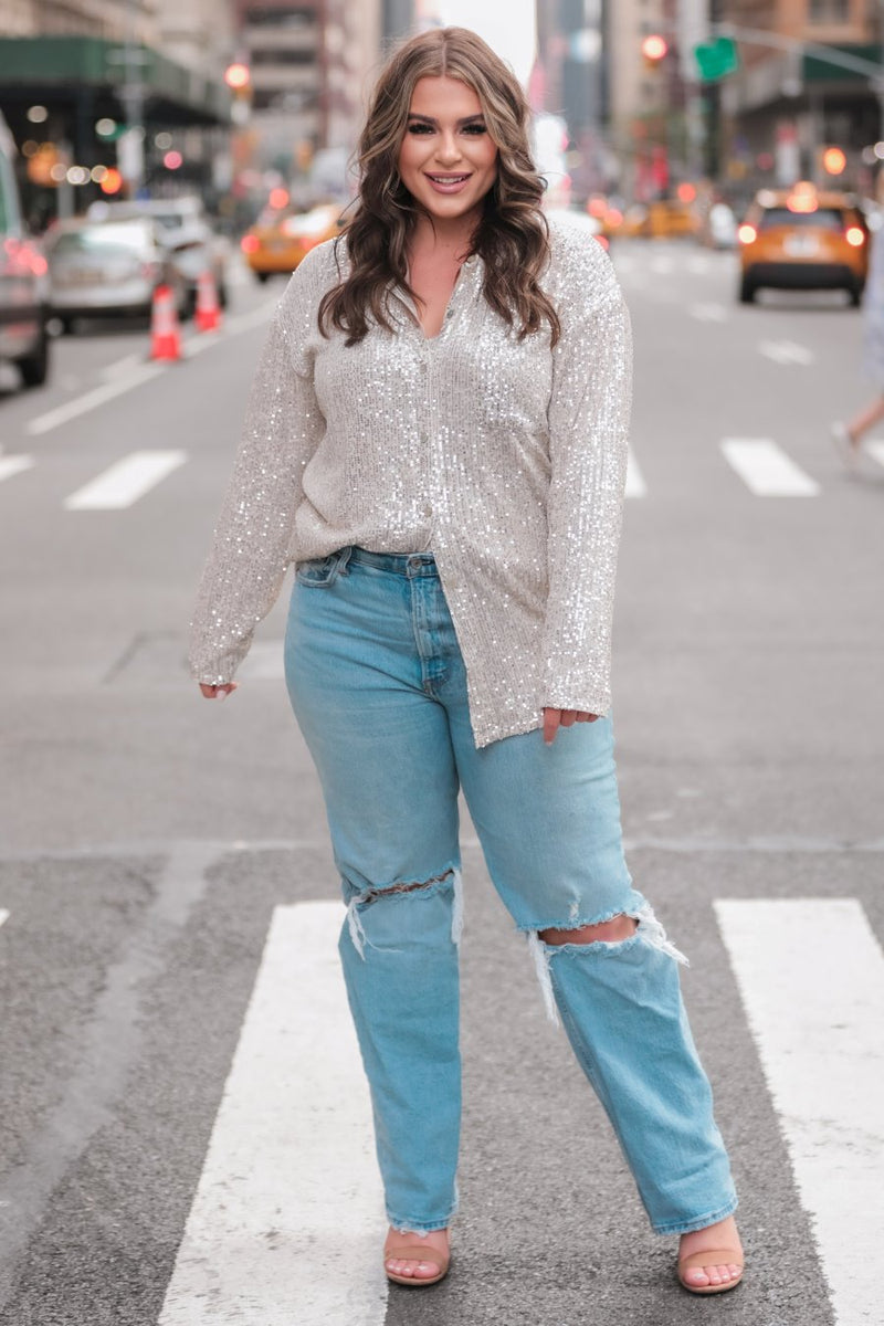 a loose long-sleeve top made of lightweight material covered in shimmering sequins with a collared neckline, fitted button closure cuffs, an accent chest pocket, and a relaxed button-up silhouette that falls into a round high-low hemline in champagne