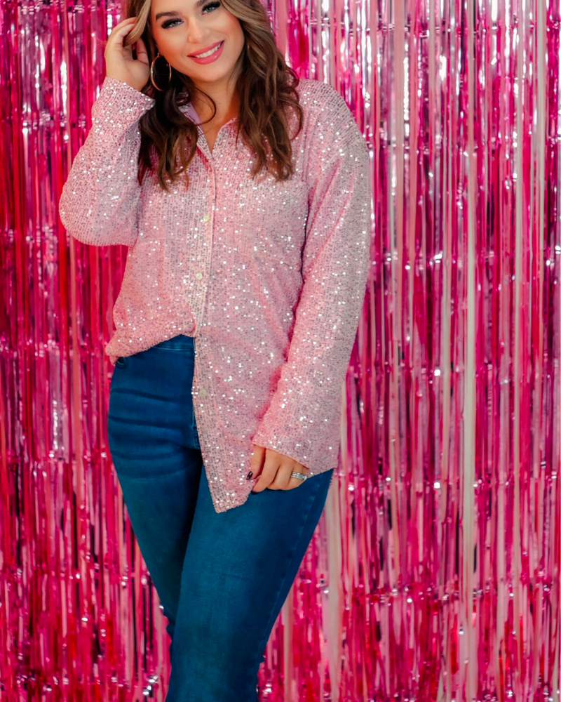 a loose long-sleeve top made of lightweight material covered in shimmering sequins with a collared neckline, fitted button closure cuffs, an accent chest pocket, and a relaxed button-up silhouette that falls into a round high-low hemline in pink