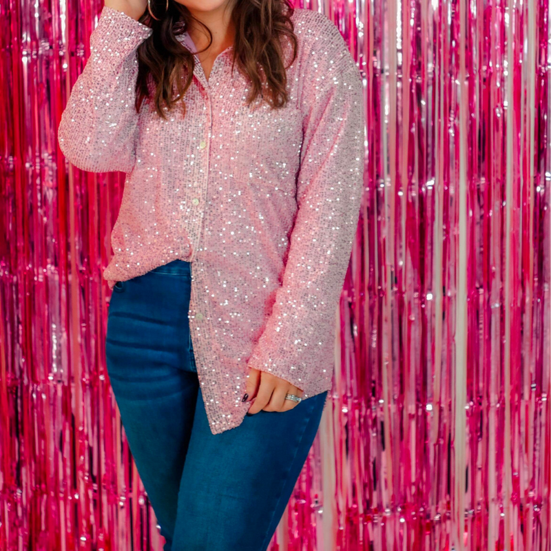 a loose long-sleeve top made of lightweight material covered in shimmering sequins with a collared neckline, fitted button closure cuffs, an accent chest pocket, and a relaxed button-up silhouette that falls into a round high-low hemline in pink