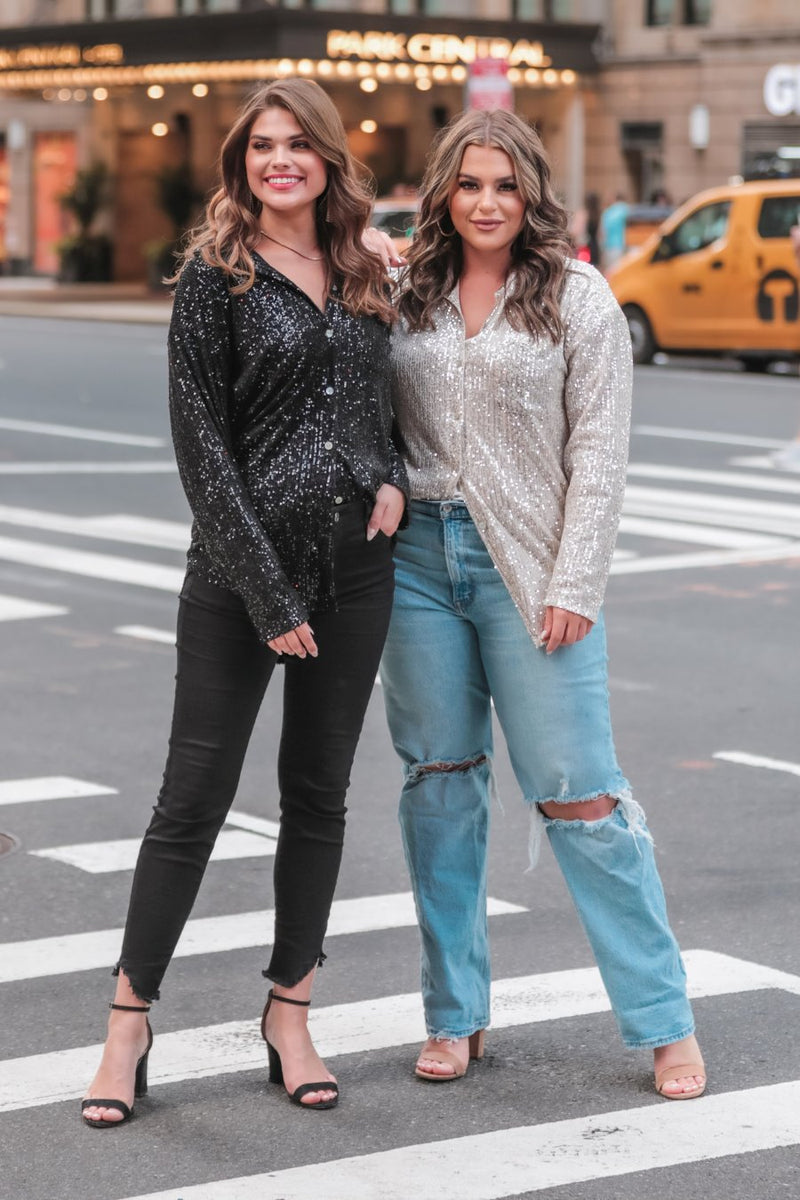 a loose long-sleeve top made of lightweight material covered in shimmering sequins with a collared neckline, fitted button closure cuffs, an accent chest pocket, and a relaxed button-up silhouette that falls into a round high-low hemline in black and champagne