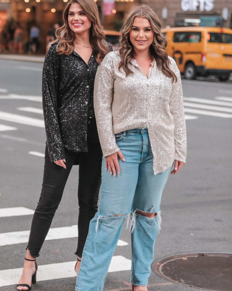 a loose long-sleeve top made of lightweight material covered in shimmering sequins with a collared neckline, fitted button closure cuffs, an accent chest pocket, and a relaxed button-up silhouette that falls into a round high-low hemline in black and champagne