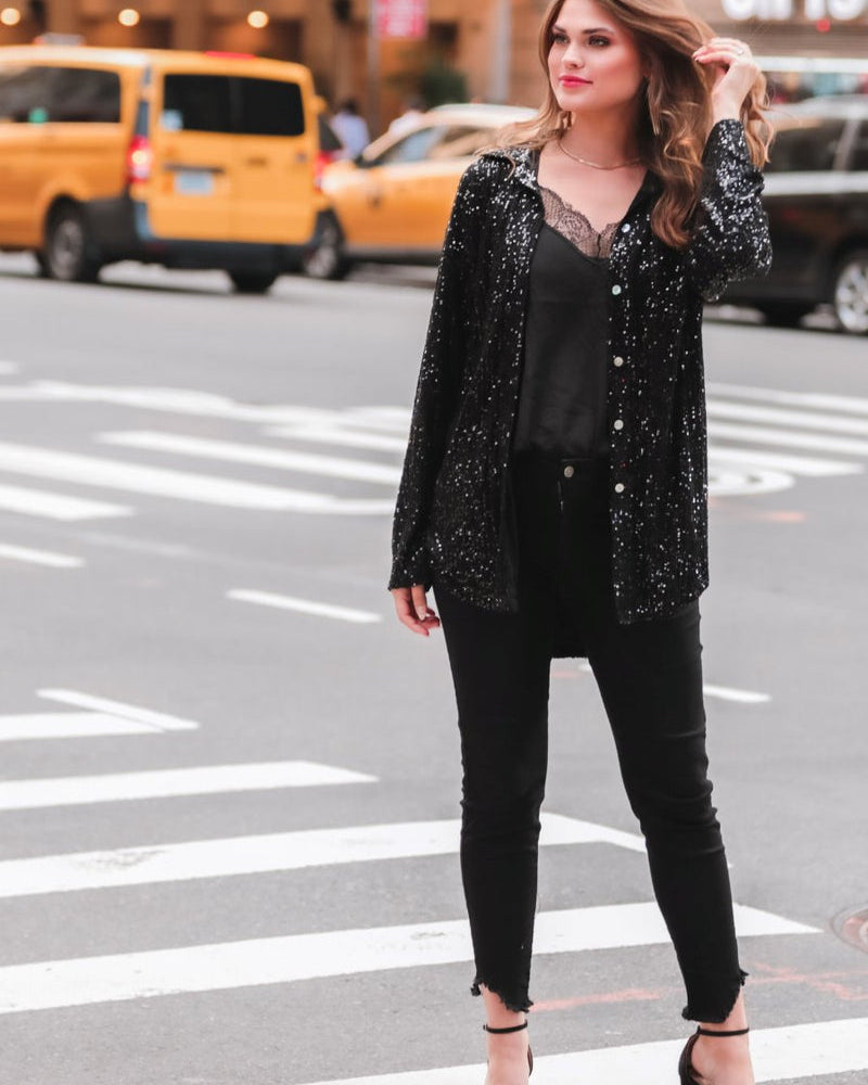 a loose long-sleeve top made of lightweight material covered in shimmering sequins with a collared neckline, fitted button closure cuffs, an accent chest pocket, and a relaxed button-up silhouette that falls into a round high-low hemline in black