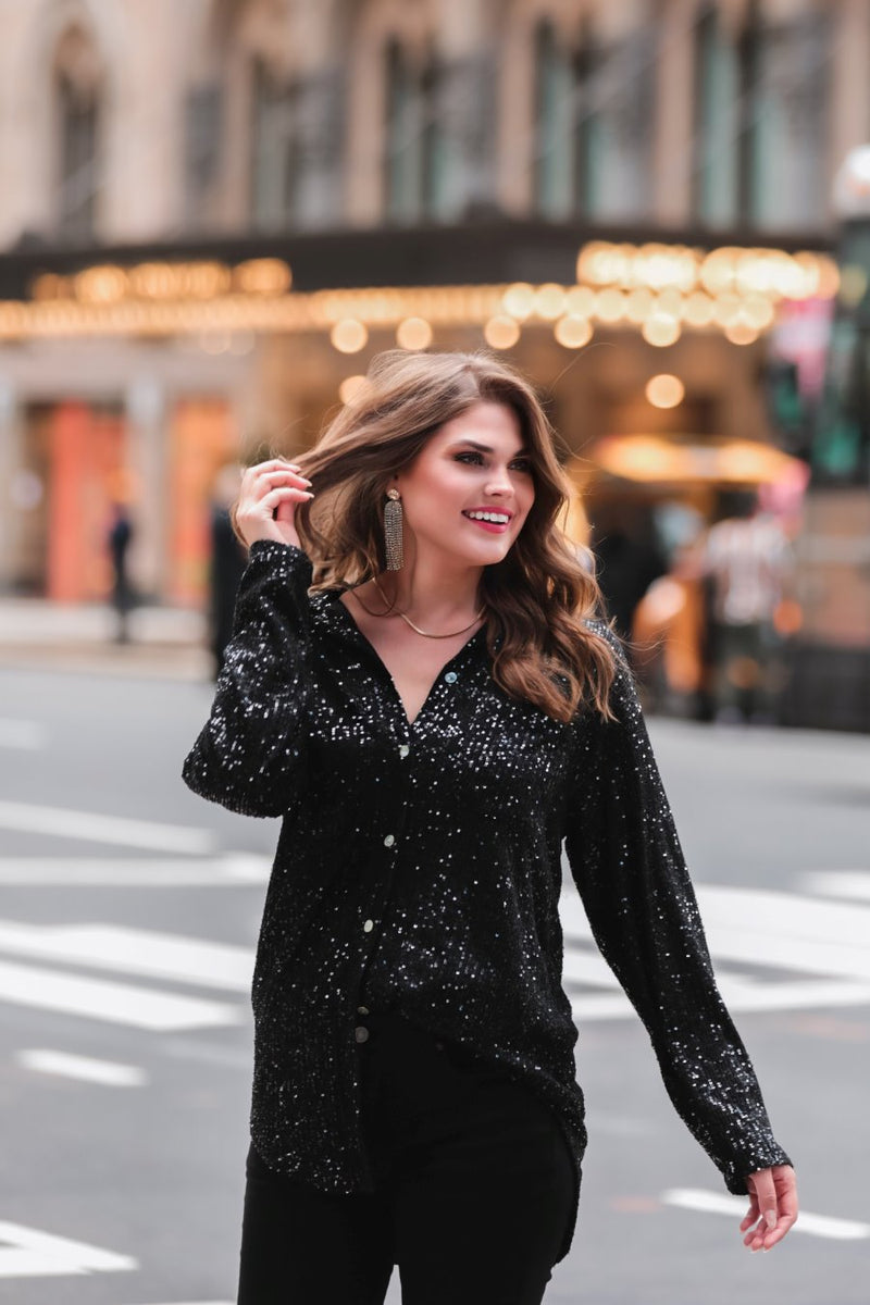 a loose long-sleeve top made of lightweight material covered in shimmering sequins with a collared neckline, fitted button closure cuffs, an accent chest pocket, and a relaxed button-up silhouette that falls into a round high-low hemline in black