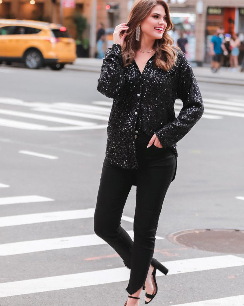 a loose long-sleeve top made of lightweight material covered in shimmering sequins with a collared neckline, fitted button closure cuffs, an accent chest pocket, and a relaxed button-up silhouette that falls into a round high-low hemline in black