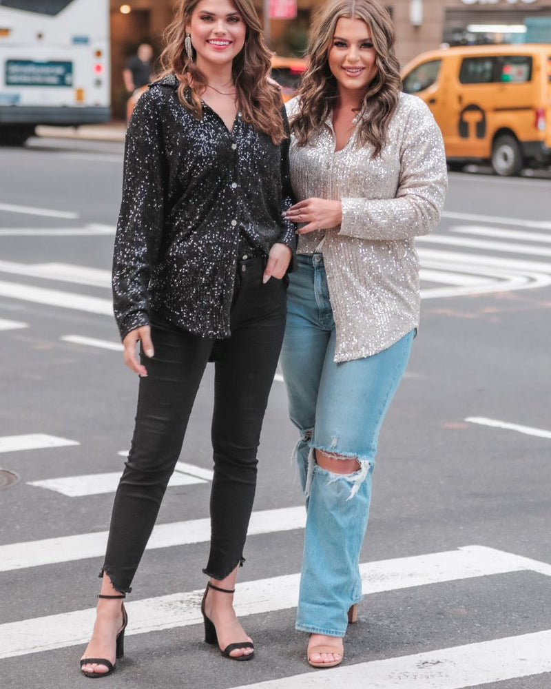 a loose long-sleeve top made of lightweight material covered in shimmering sequins with a collared neckline, fitted button closure cuffs, an accent chest pocket, and a relaxed button-up silhouette that falls into a round high-low hemline in black and champagne