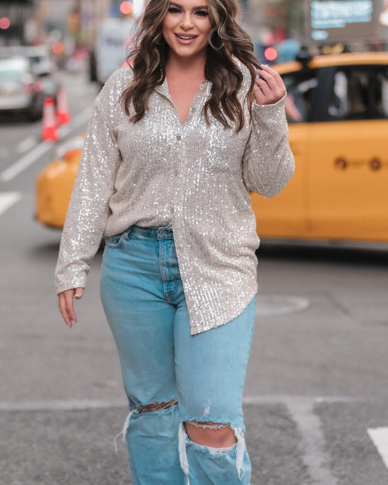 a loose long-sleeve top made of lightweight material covered in shimmering sequins with a collared neckline, fitted button closure cuffs, an accent chest pocket, and a relaxed button-up silhouette that falls into a round high-low hemline in champagne