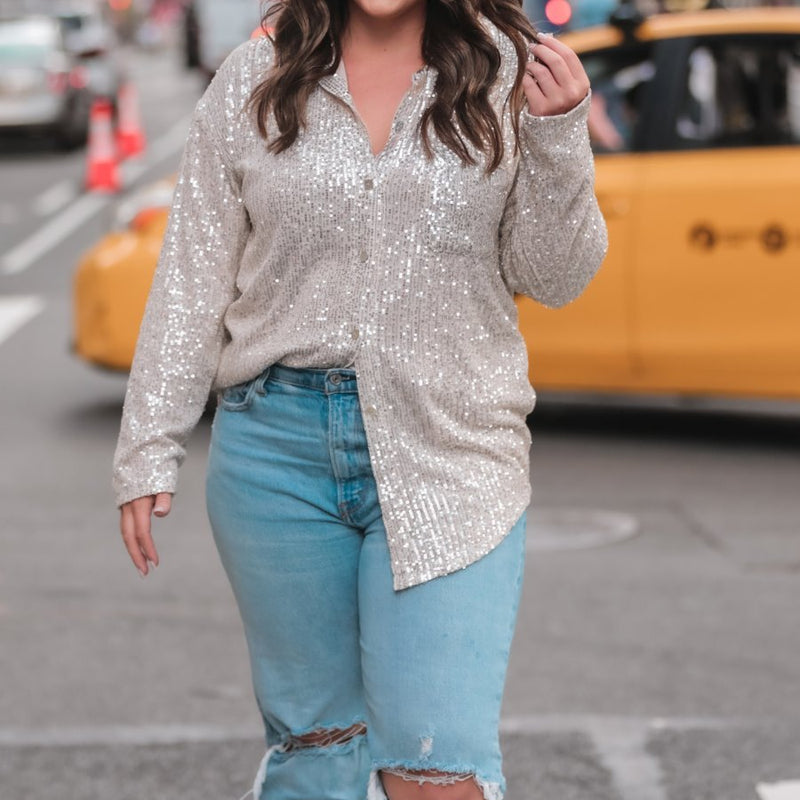 a loose long-sleeve top made of lightweight material covered in shimmering sequins with a collared neckline, fitted button closure cuffs, an accent chest pocket, and a relaxed button-up silhouette that falls into a round high-low hemline in champagne