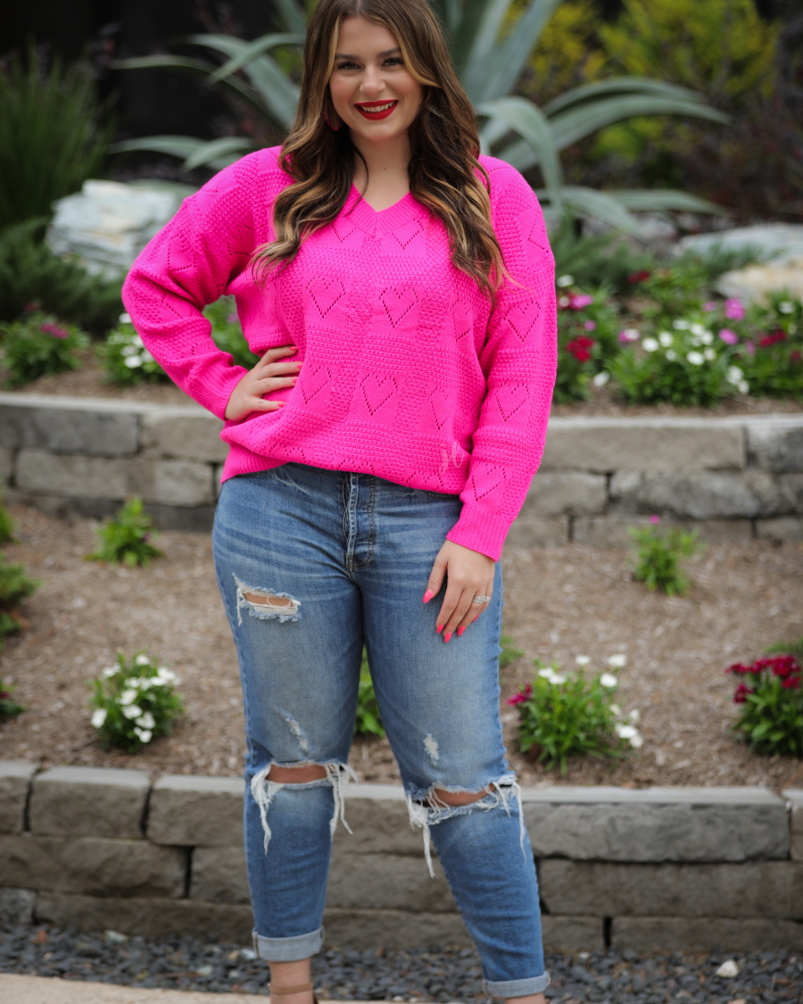 a pink long-sleeve open-knit sweater with a cutout heart pattern and ribbed detailing on the V-neckline, cuffs, and hemline