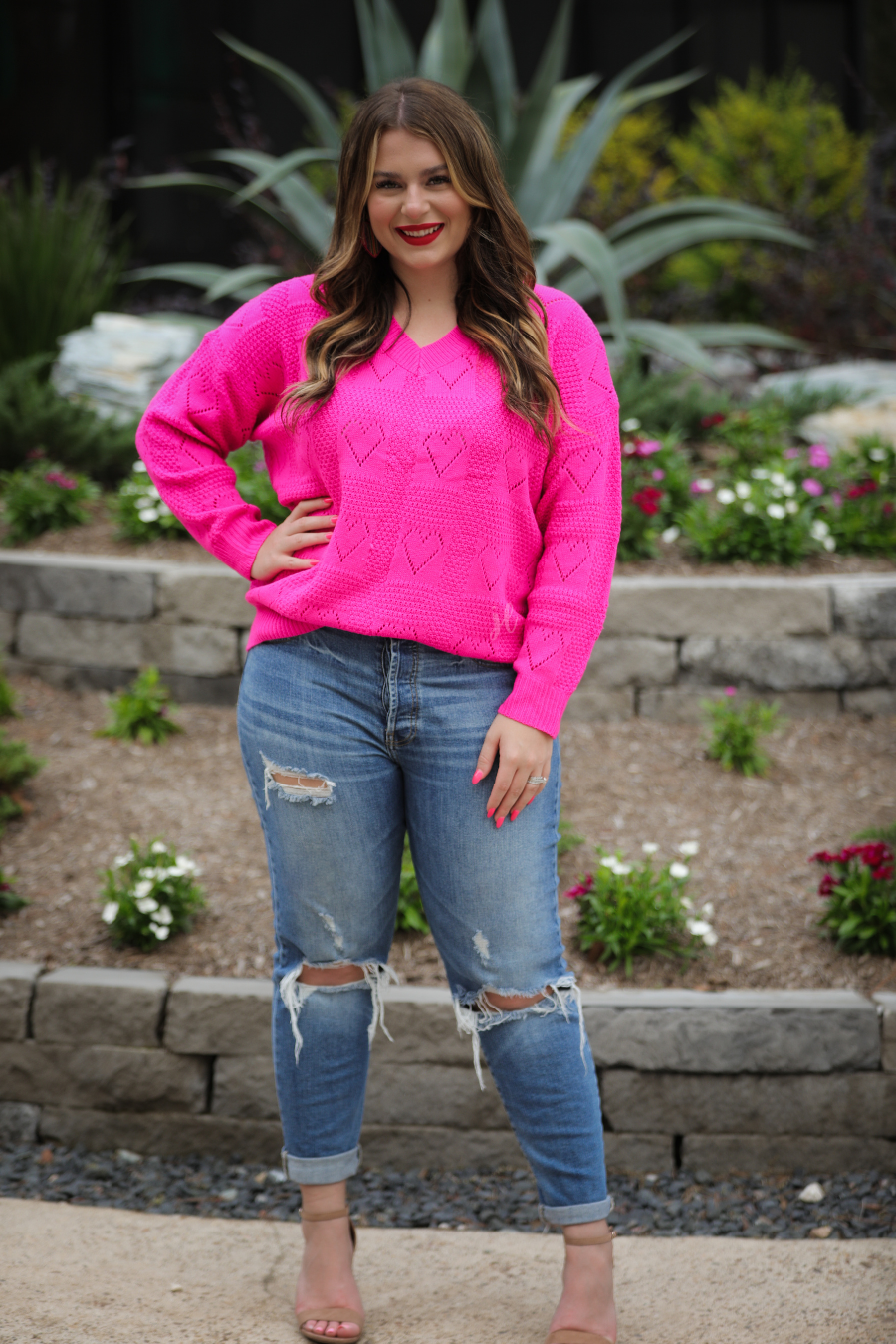 a pink long-sleeve open-knit sweater with a cutout heart pattern and ribbed detailing on the V-neckline, cuffs, and hemline