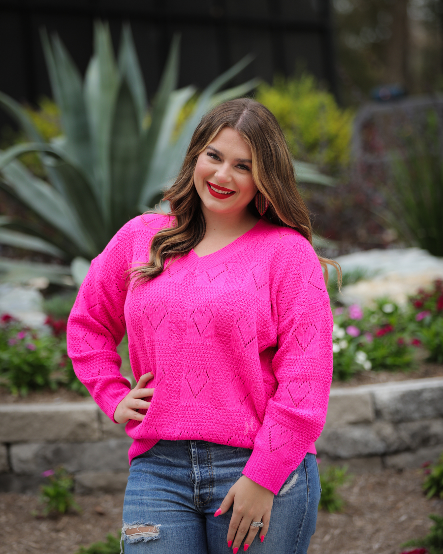 a pink long-sleeve open-knit sweater with a cutout heart pattern and ribbed detailing on the V-neckline, cuffs, and hemline