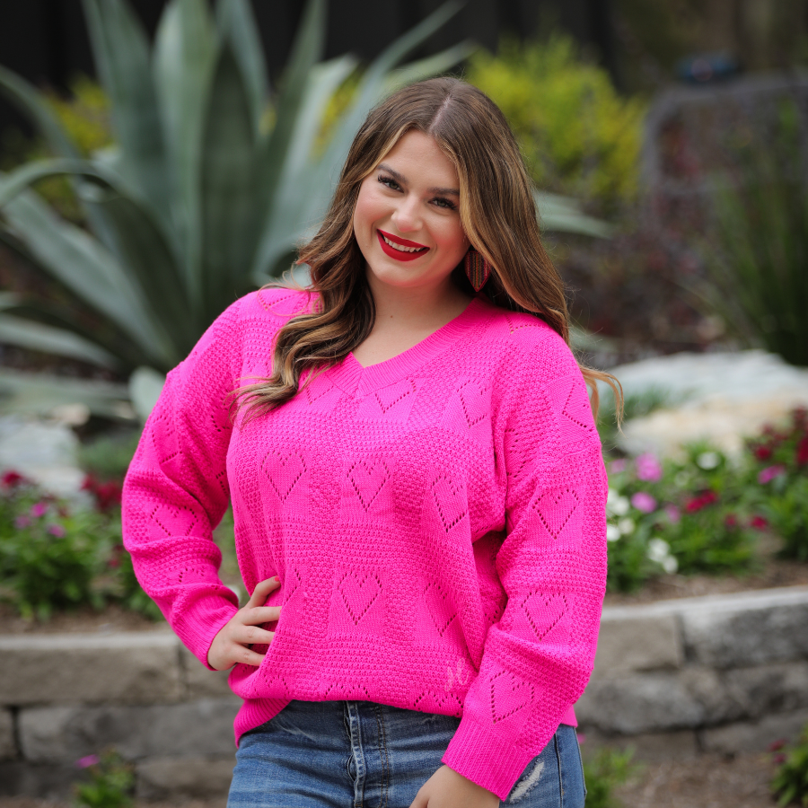 a pink long-sleeve open-knit sweater with a cutout heart pattern and ribbed detailing on the V-neckline, cuffs, and hemline