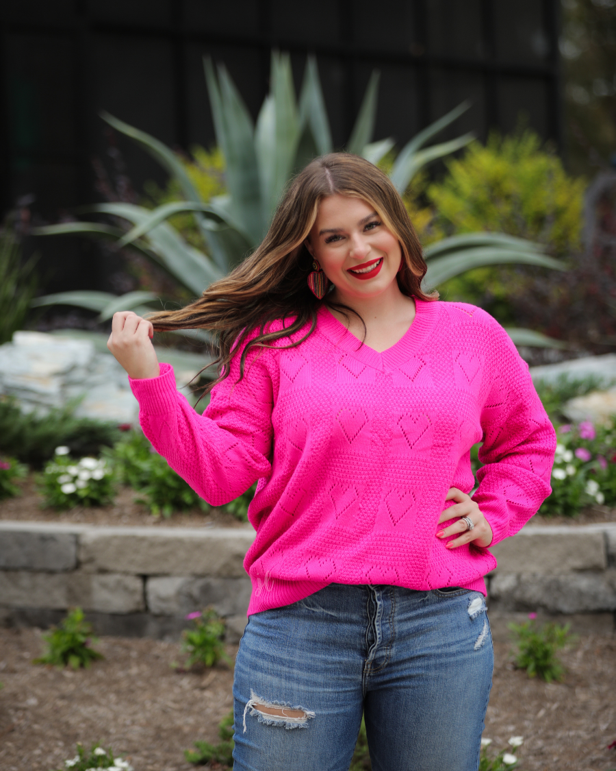 a pink long-sleeve open-knit sweater with a cutout heart pattern and ribbed detailing on the V-neckline, cuffs, and hemline